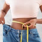 A woman holding a tape measure around her waist, wearing oversized jeans, symbolizing weight loss and transformation.
