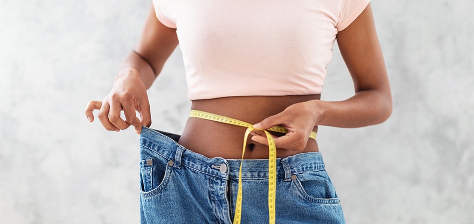 A woman holding a tape measure around her waist, wearing oversized jeans, symbolizing weight loss and transformation.